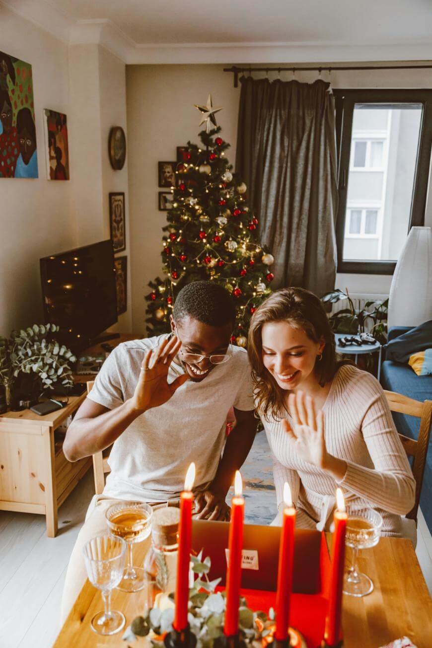 A smiling couple sits at a table with red candles, waving at a laptop screen during a video call. A decorated Christmas tree with lights and ornaments stands behind them in a cozy, festive living room.