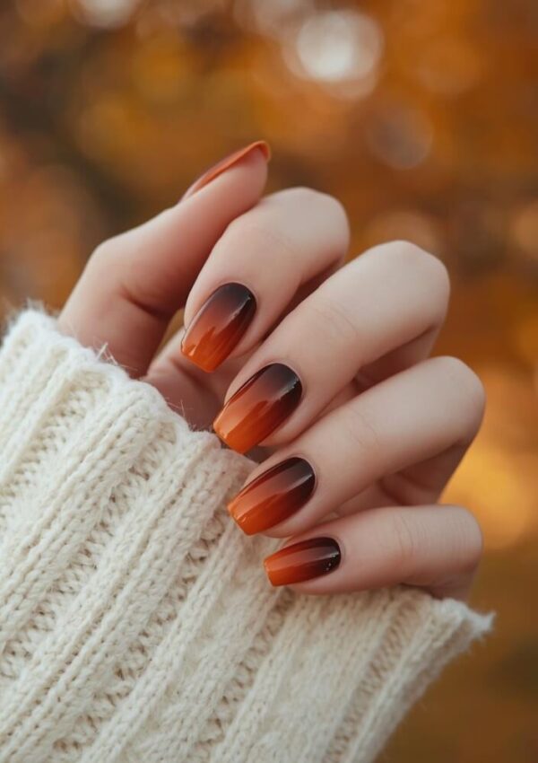 A hand with long, almond-shaped nails featuring a brown and orange ombre manicure is shown against a blurred autumn background. The person is wearing a cozy white knitted sweater.