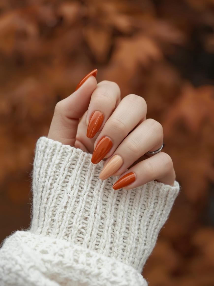A hand with long, almond-shaped nails painted in autumn shades of orange and nude, wearing a cozy white knit sweater, is held up against a blurred background of fall-colored leaves.