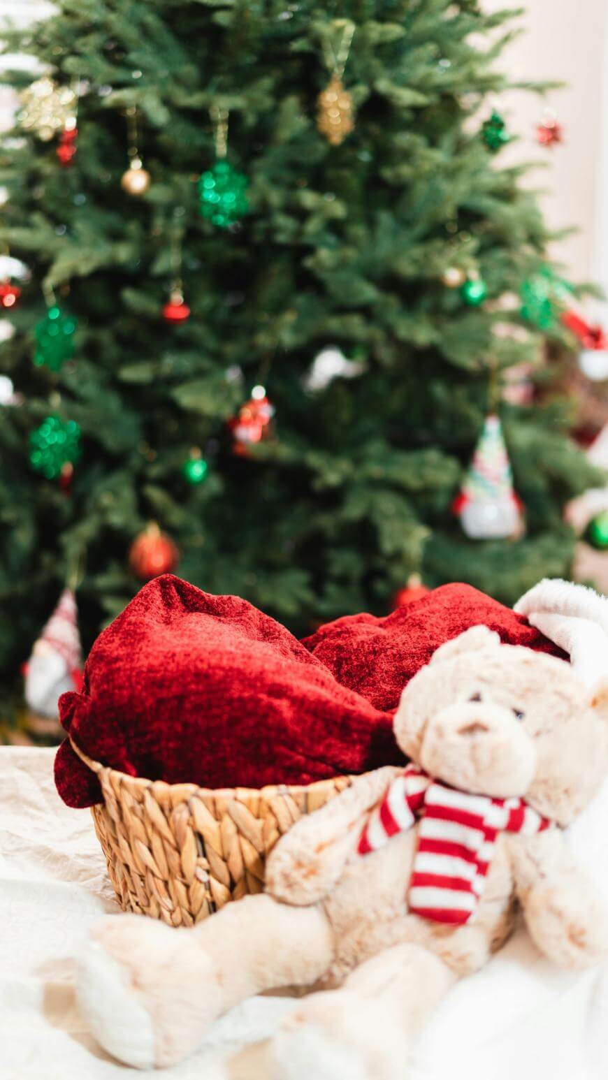 A wicker basket with a red blanket sits in front of a decorated Christmas tree. A plush teddy bear with a red and white striped scarf lies beside the basket on a white surface.