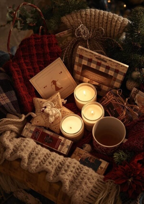 A cozy holiday gift basket filled with candles, a hot drink, knitted blankets, festive cards, and wrapped gifts, surrounded by greenery and red flowers, creating a warm and inviting Christmas atmosphere.