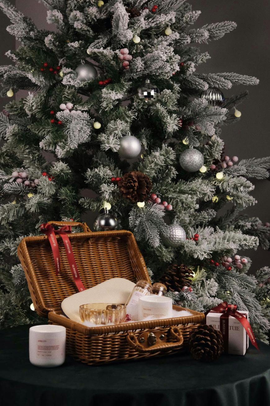 A wicker picnic basket with candles, glass cups, and wrapped gifts sits on a table in front of a decorated, snow-dusted Christmas tree adorned with silver ornaments and pine cones.