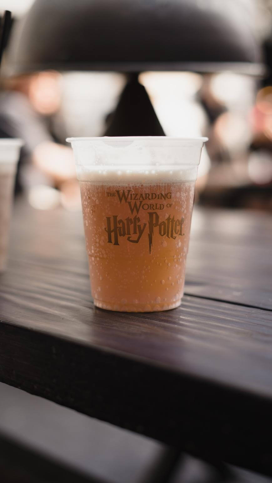 A plastic cup filled with a fizzy, amber-colored drink sits on a dark wooden table. The cup is labeled The Wizarding World of Harry Potter. The background is blurred with indistinct shapes and people.