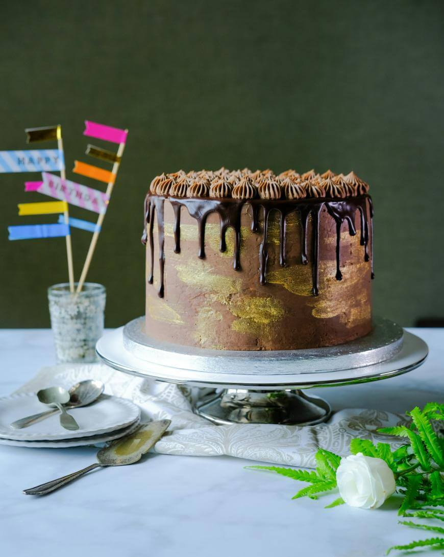 A chocolate drip cake with gold accents sits on a silver stand. The cake is topped with piped frosting. Nearby are birthday flag decorations in a glass, cutlery, and a white rose on a marble surface.