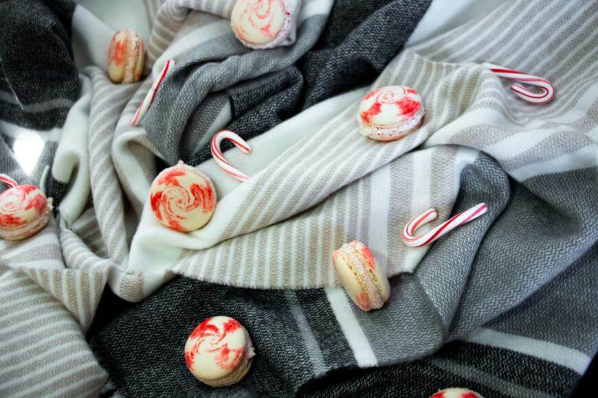 Red and white swirled macarons and mini candy canes are scattered on a soft, striped gray and white fabric, creating a festive and cozy arrangement.