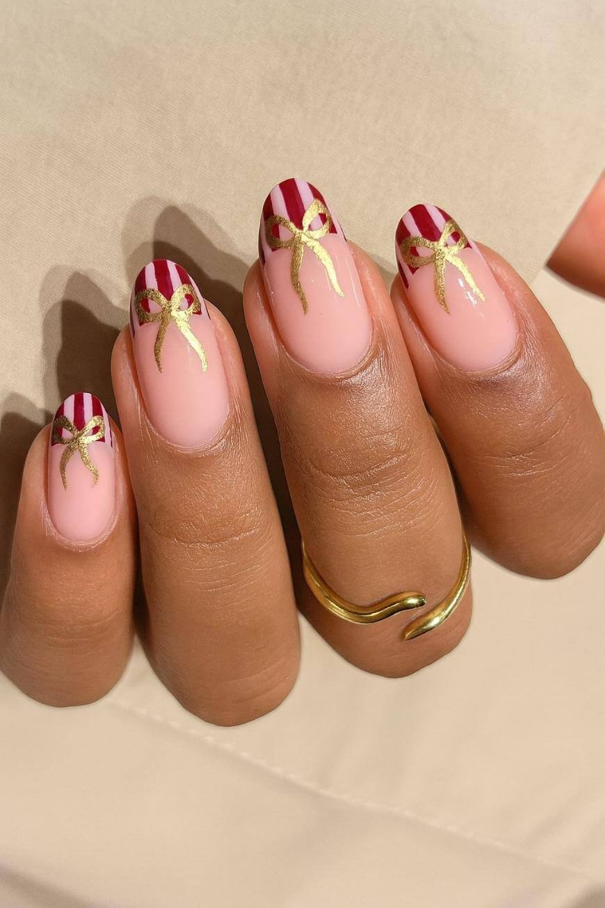A hand with four fingernails painted with red and white stripes at the tips, each decorated with a gold bow design. The base of the nails is a natural nude color. The person wears a gold wavy ring.