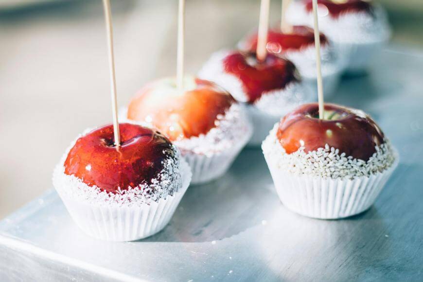 Four shiny red candy apples with wooden sticks, dipped in coconut flakes around the base, are placed in white paper cups on a metallic surface.
