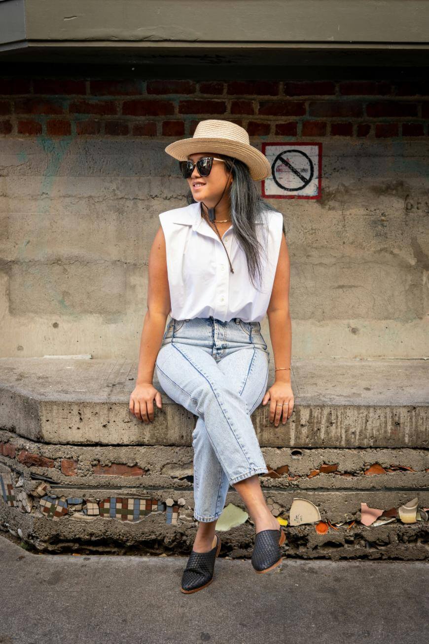 A woman in a sleeveless white shirt, light blue jeans, black shoes, sunglasses, and a straw hat sits on a concrete bench against a brick wall with a no smoking sign behind her.