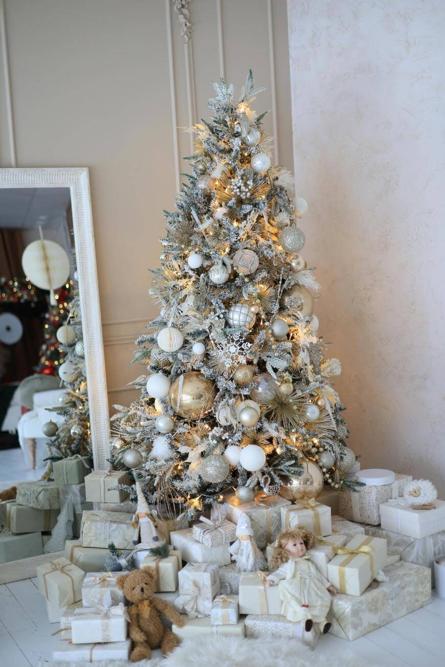 A Christmas tree decorated with white and gold ornaments stands in a bright room. Numerous wrapped gifts, teddy bears, and dolls are placed at its base, and a large mirror reflects part of the festive scene.
