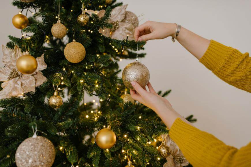 A person wearing a yellow sweater decorates a Christmas tree with gold baubles and fairy lights. The tree also features gold poinsettia ornaments.