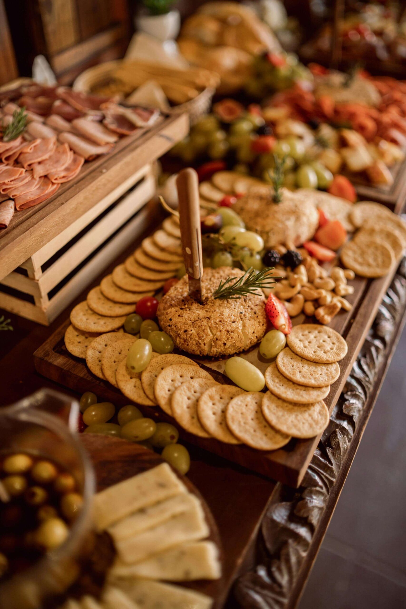 A charcuterie board with round cheese balls topped with rosemary, surrounded by crackers, grapes, nuts, and sliced strawberries. Nearby are sliced deli meats, cheese, and a bowl of olives.