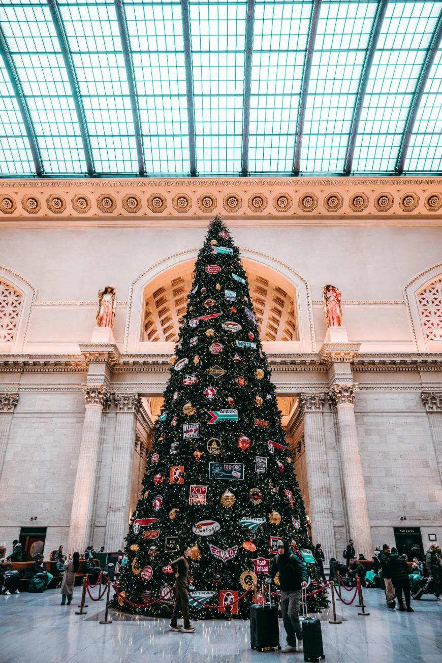 A large, decorated Christmas tree stands in the center of a grand train station with high arched ceilings and a glass roof. People are gathered around the tree, some with suitcases.