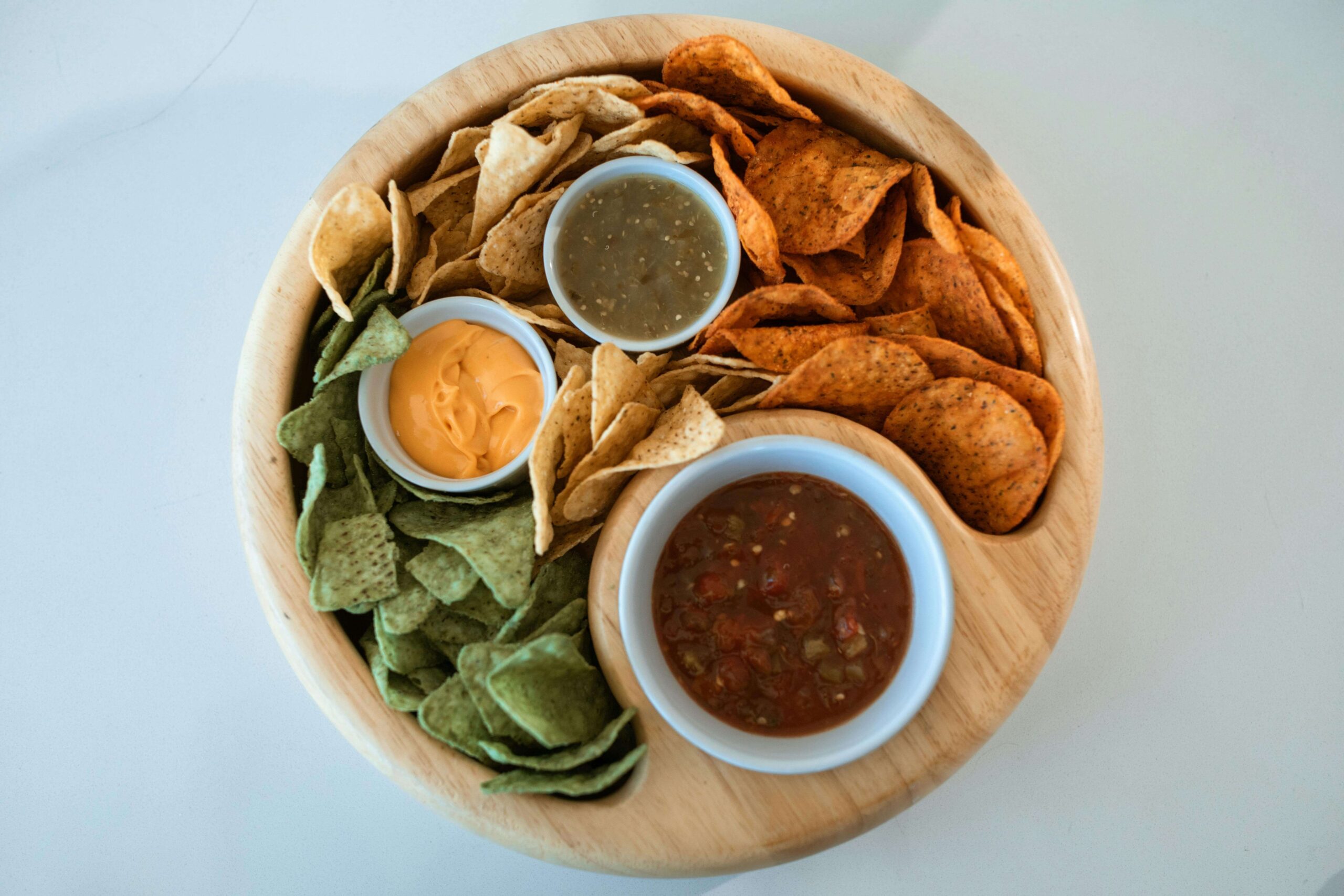A wooden platter holds three kinds of chips—green, yellow, and red—arranged around three dips: yellow cheese, red salsa, and green salsa, each in a small white bowl.