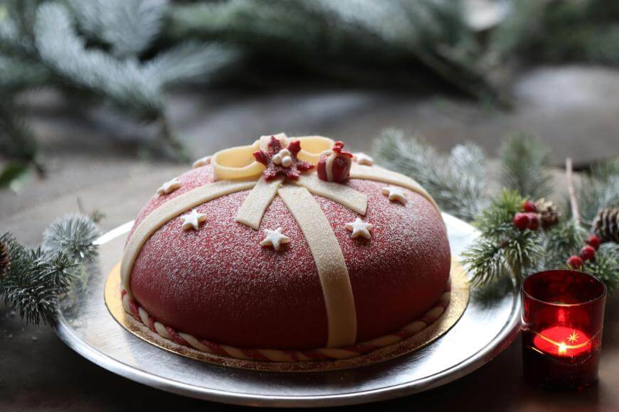 A festive round cake decorated with red icing, white chocolate ribbons, stars, and small holiday-themed toppers, dusted with powdered sugar. Pine branches and a lit red candle surround the cake.