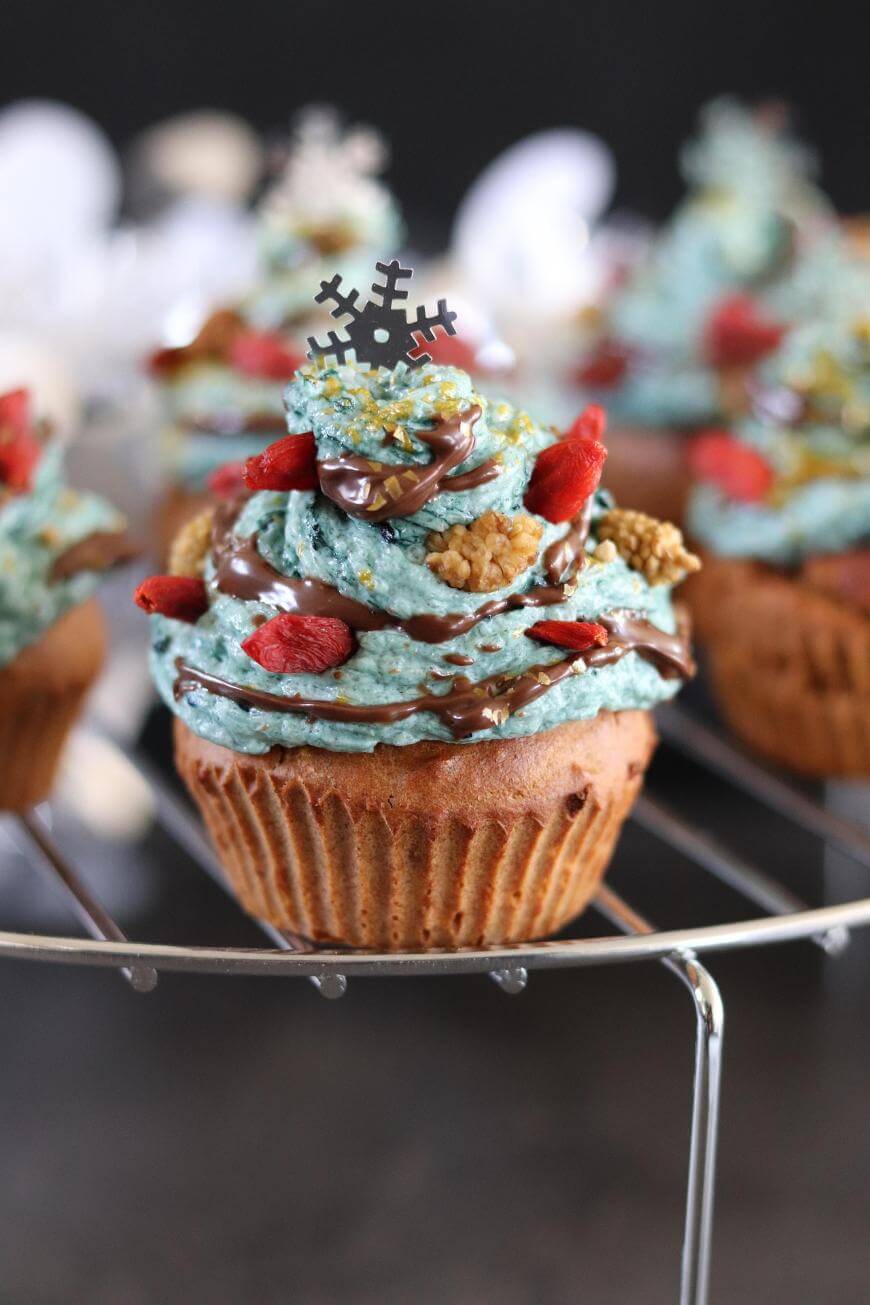 A cupcake with blue frosting, topped with a chocolate drizzle, dried berries, and a decorative snowflake topper, displayed on a wire rack. Other decorated cupcakes are blurred in the background.