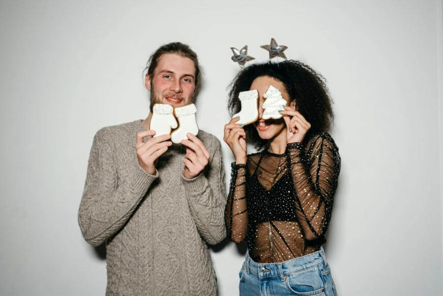 A man and a woman stand against a white background, smiling and holding Christmas-themed cookies. The woman wears star headbands and a sheer black top, holding cookies in front of her face. The man wears a gray sweater.