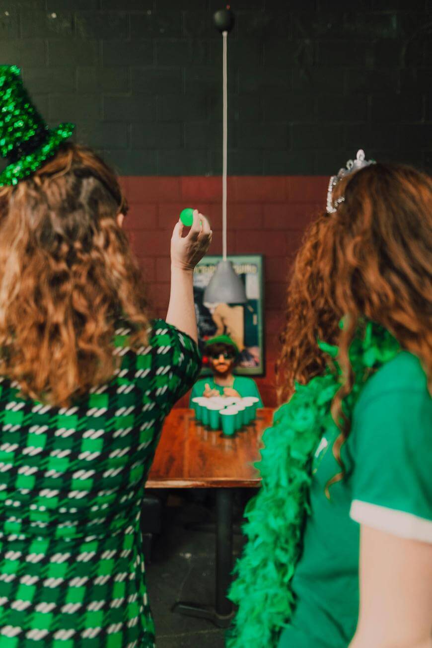 Two women in festive green outfits face a table, one aiming to throw a ping pong ball during a game of beer pong. The background is a dimly lit room decorated for a celebration.