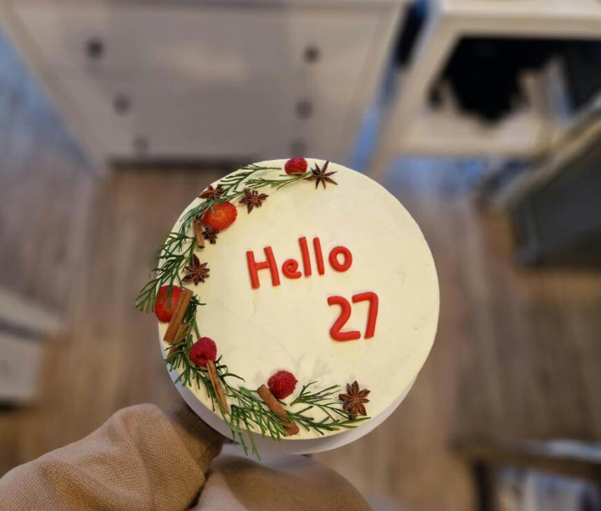 A round white cake with “Hello 27” written in red icing. The cake is decorated with pine leaves, cinnamon sticks, and red berries on one side. The cake is being held in a hand above a wooden floor.