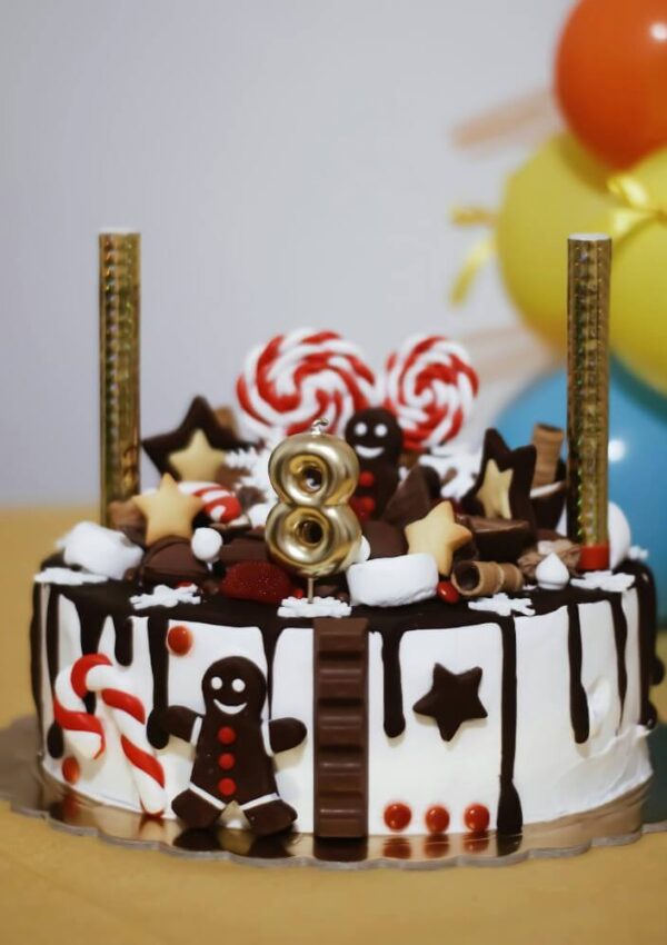 A decorated birthday cake with chocolate drizzle, gingerbread men, candy canes, lollipops, star-shaped cookies, and a gold number 8 candle. Colorful balloons are in the background.