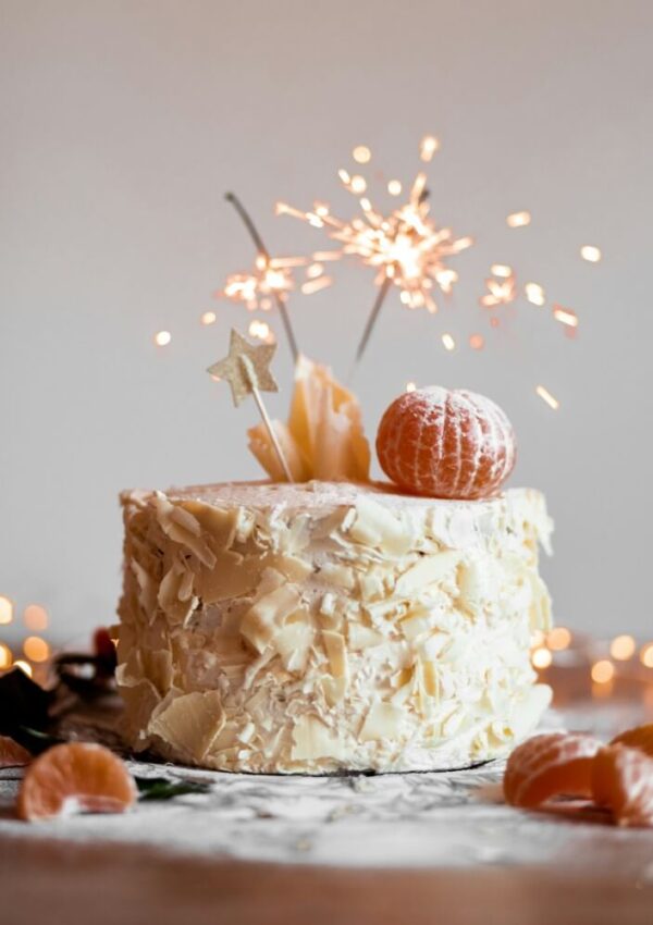 A festive cake decorated with white chocolate shavings, a peeled mandarin, star toppers, and sparklers, surrounded by mandarins and dusted with powdered sugar, with fairy lights in the background.