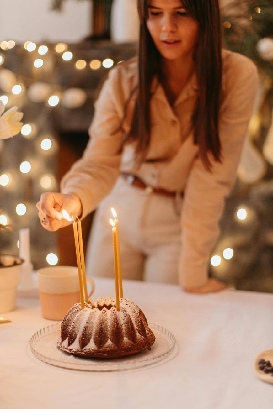 A woman in beige clothes lights tall candles on a bundt cake dusted with powdered sugar, with warm string lights and festive decorations in the background.