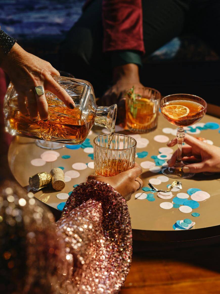 A person pours whiskey from a decanter into a glass on a table decorated with confetti, corks, and three cocktails, while others reach for their drinks.