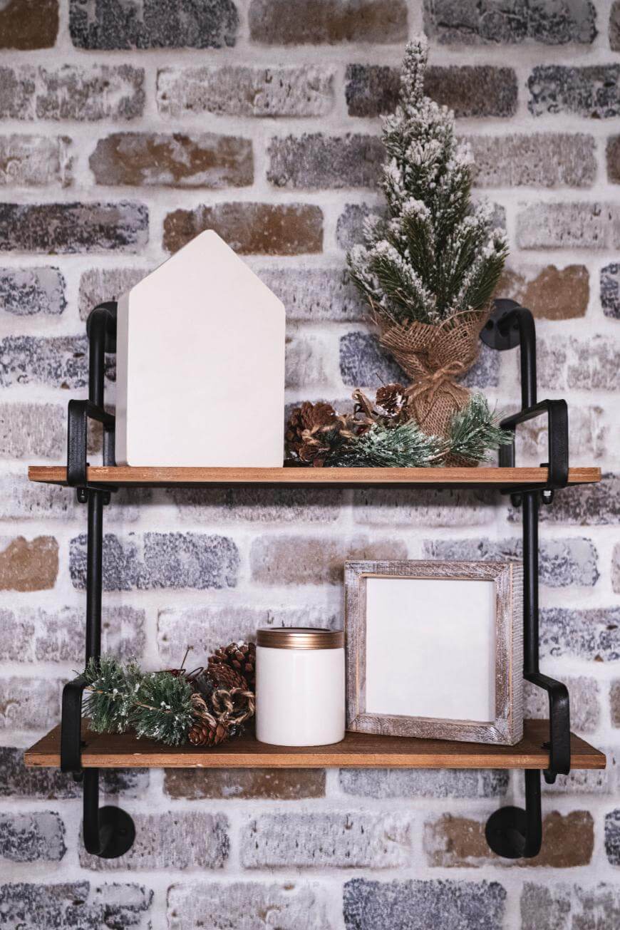 Two wooden shelves on a brick wall display a small potted pine tree, a white house-shaped ornament, pinecones, a white candle, greenery, and an empty picture frame for a rustic, winter-themed decor.