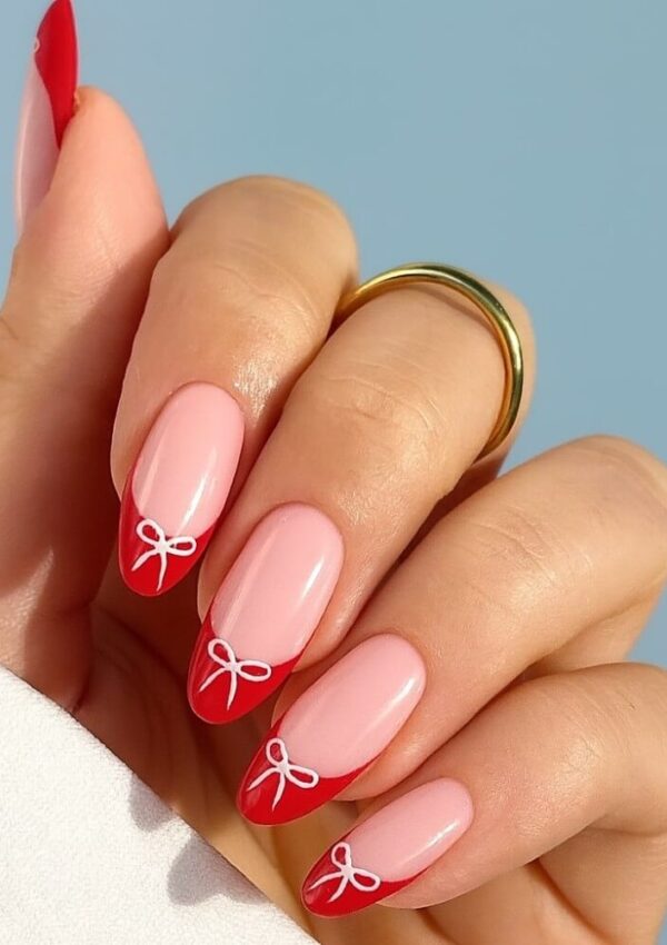 A hand with long almond-shaped nails featuring a pink base and red tips, each tip decorated with a small white bow design. The person is wearing a gold ring on their middle finger.
