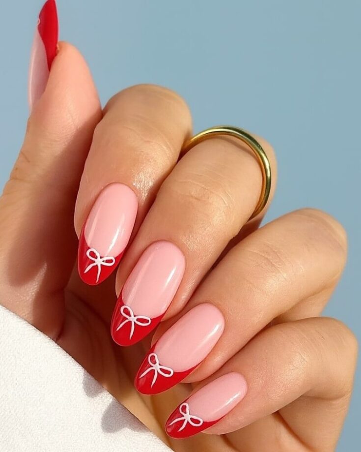 A hand with long almond-shaped nails featuring a pink base and red tips, each tip decorated with a small white bow design. The person is wearing a gold ring on their middle finger.