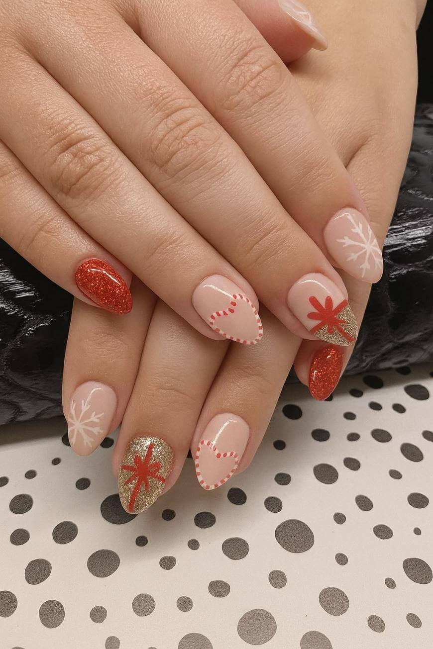 A close-up of hands with festive nail art featuring red glitter, gold polish, white snowflakes, red stitched hearts, and red starburst designs on a polka-dotted surface.