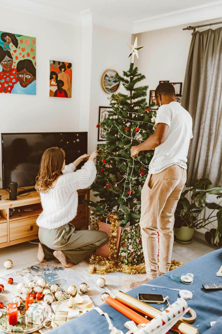 A woman and man decorate a Christmas tree with ornaments and lights in a cozy living room. Decorations and wrapping supplies are scattered on the floor, and wrapped gifts are placed under the tree.