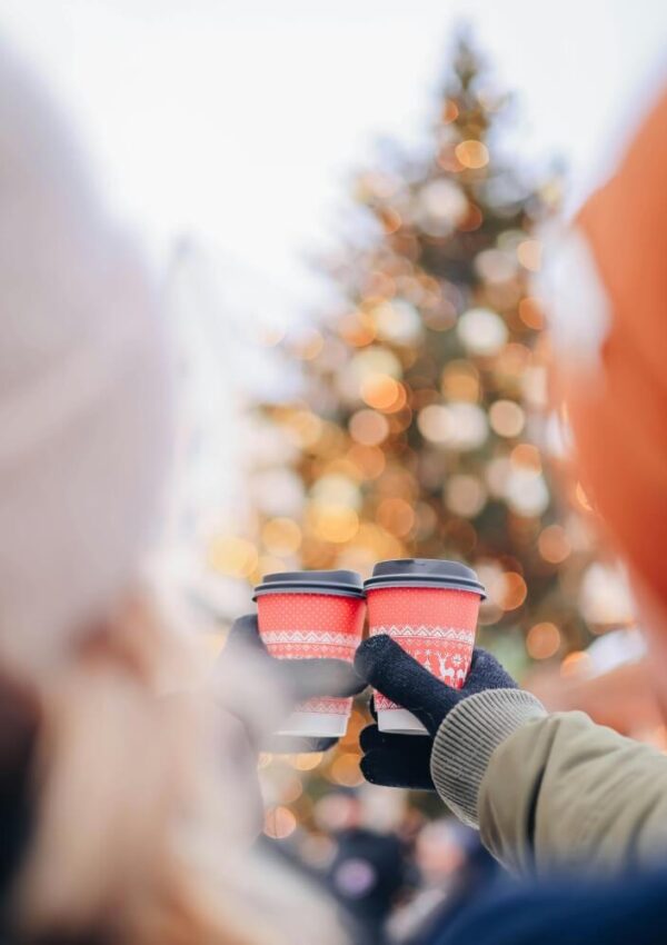 Two people wearing winter clothes hold red coffee cups in front of a decorated, blurred Christmas tree outdoors, celebrating together in a festive atmosphere.