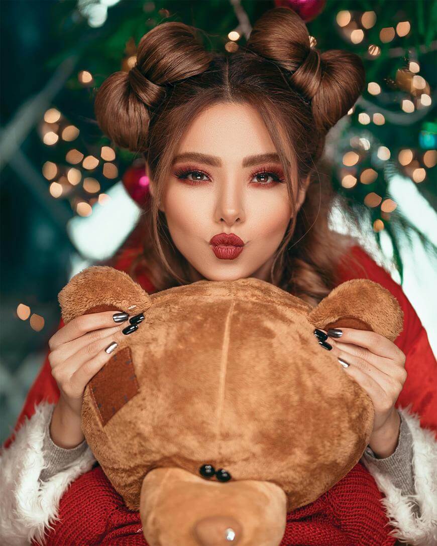 A woman with festive makeup and two space buns poses playfully, holding a plush teddy bear in front of her. She wears a red outfit, with blurred Christmas lights in the background.