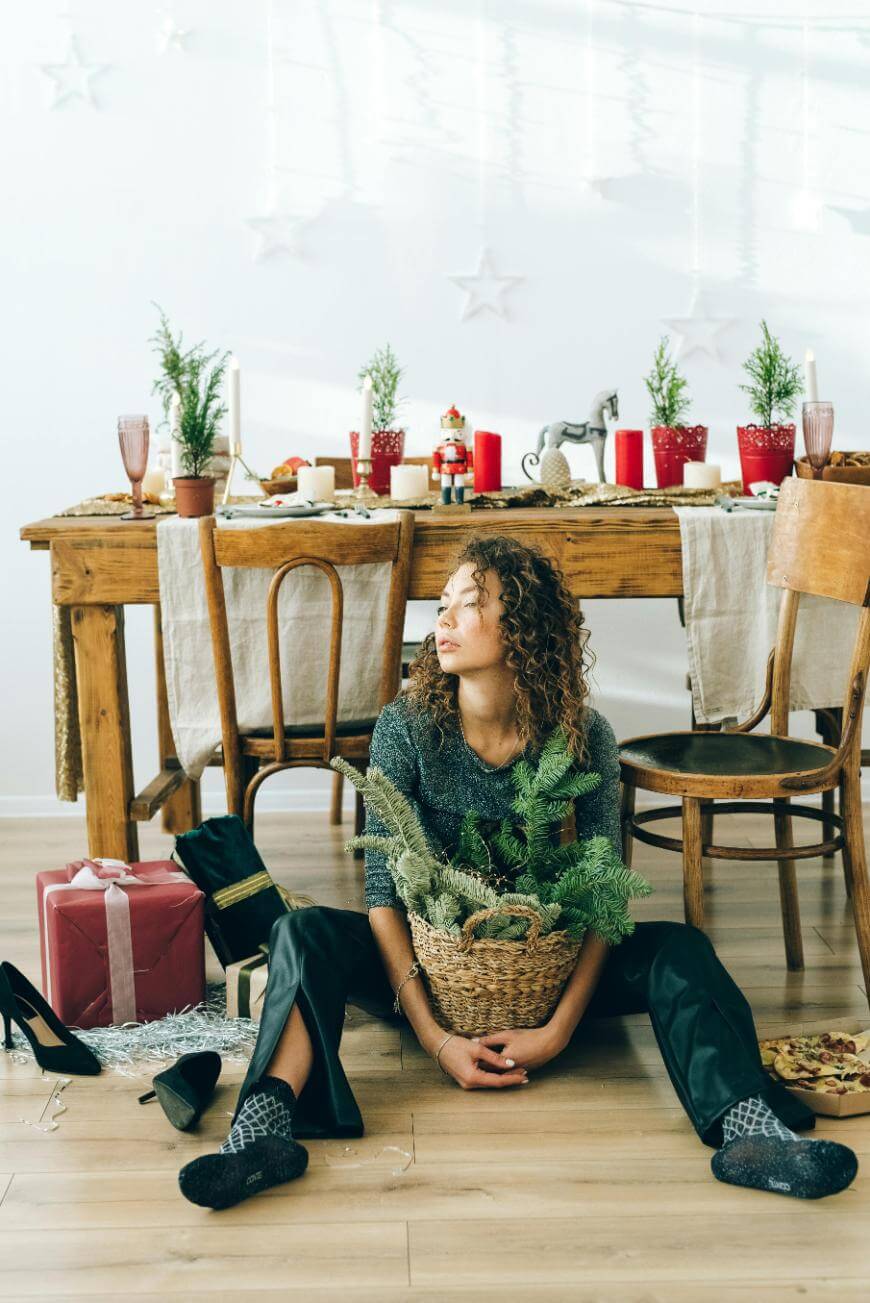 A woman with curly hair sits on the floor holding a basket of pine branches. Behind her is a festive table with holiday decorations, red cups, wrapped gifts, and party items scattered around.