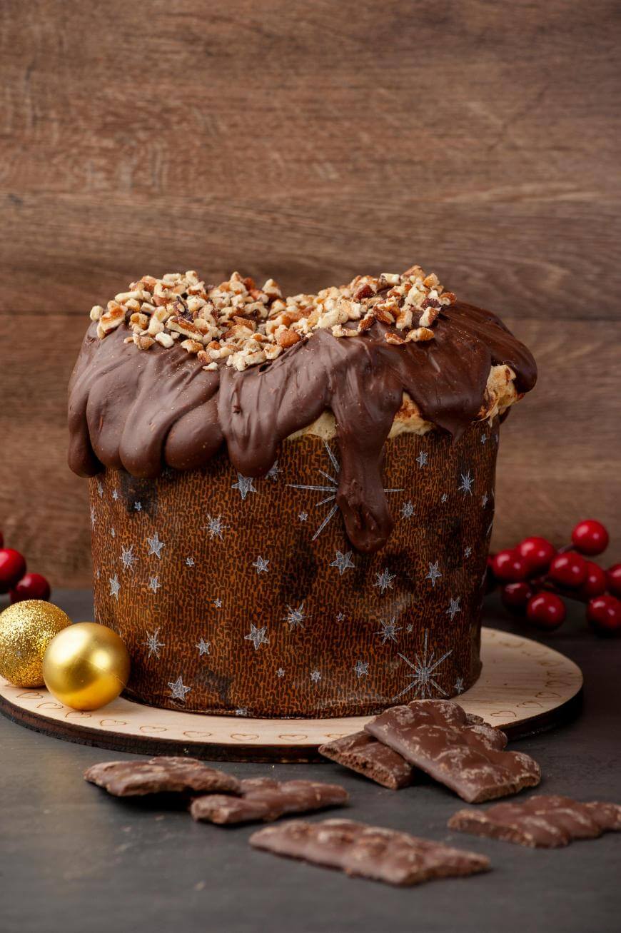 A festive panettone covered in chocolate and chopped nuts sits on a wooden board, surrounded by chocolate pieces, red berries, and gold Christmas ornaments against a rustic wooden background.