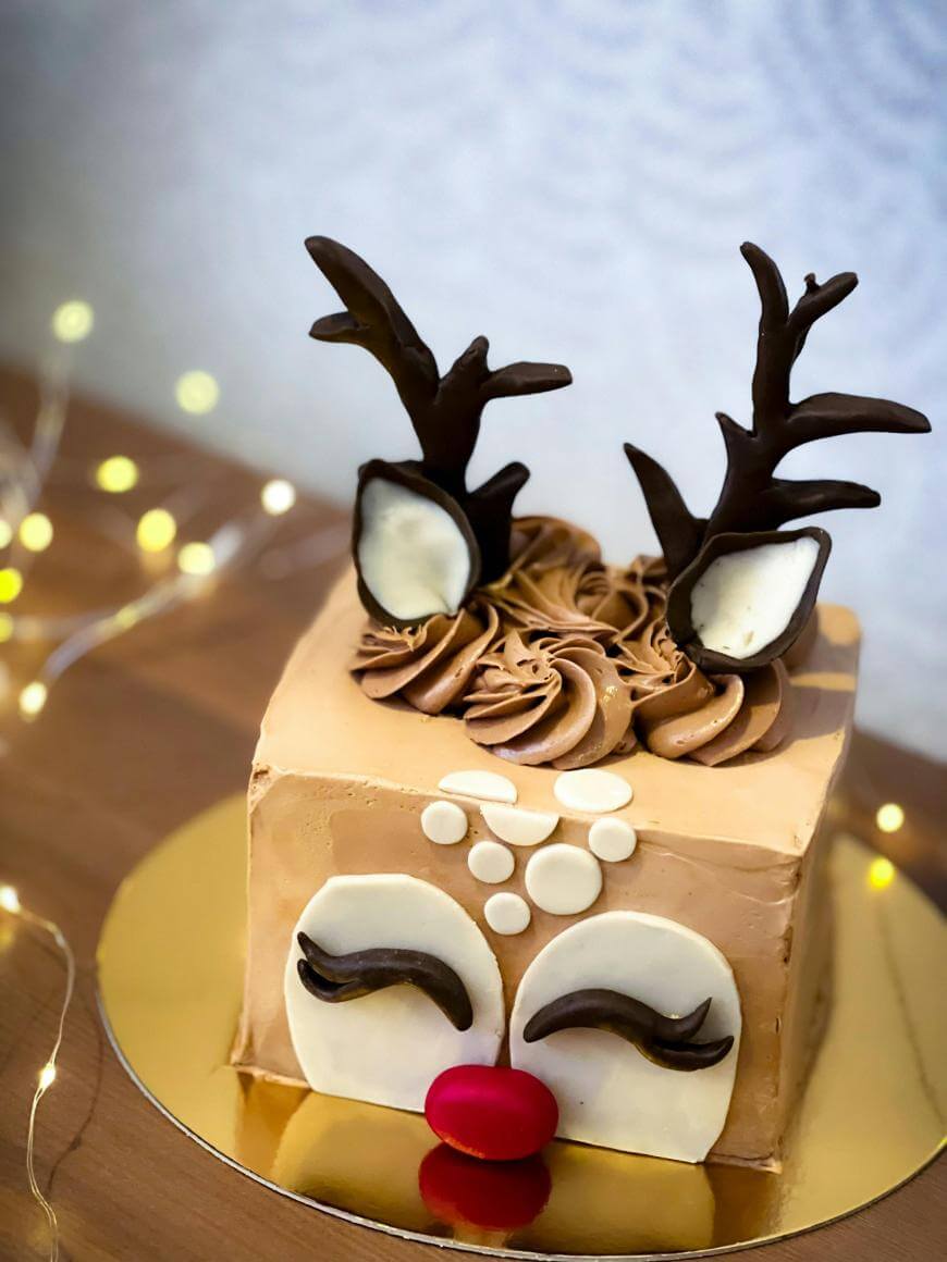 A square cake decorated as a reindeer with chocolate antlers, white ears, closed eyes with long lashes, a red nose, and beige icing, set on a gold board with string lights in the background.