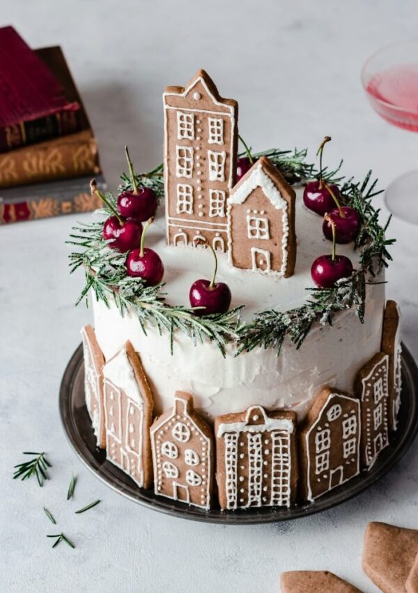 A round cake decorated with white frosting, gingerbread cookies shaped like houses, sugared rosemary, and fresh cherries, with a few vintage books and a pink drink in the background.