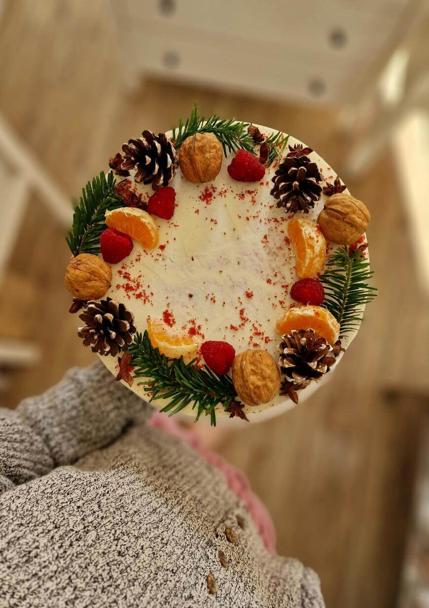 A round cake topped with white frosting, decorated with mandarin orange slices, walnuts, pinecones, strawberries, and green pine leaves, held by a person wearing a gray sweater.