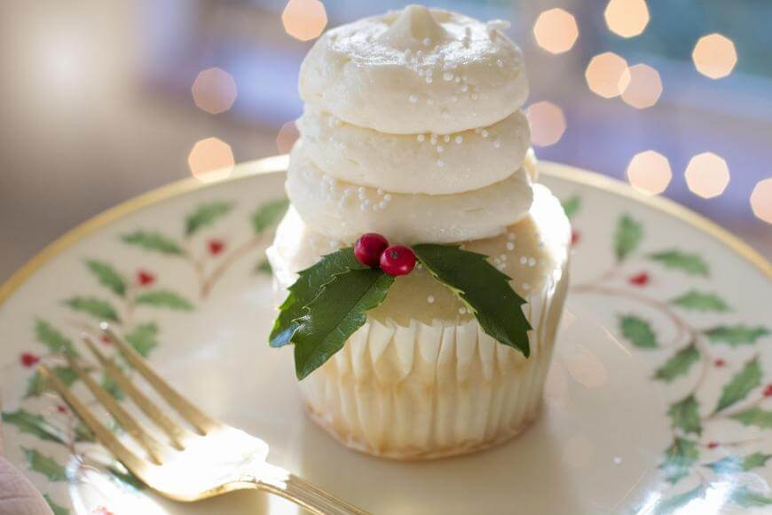 A cupcake with swirled white frosting, sprinkled with white sugar pearls, decorated with holly leaves and red berries, sits on a festive plate with a gold fork beside it. Blurred lights are in the background.