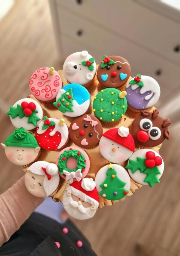 A hand holds a tray of decorated Christmas cookies shaped like holiday characters, wreaths, and festive designs, including Santa, elves, reindeer, snowmen, and holly, all colorful and intricately detailed.