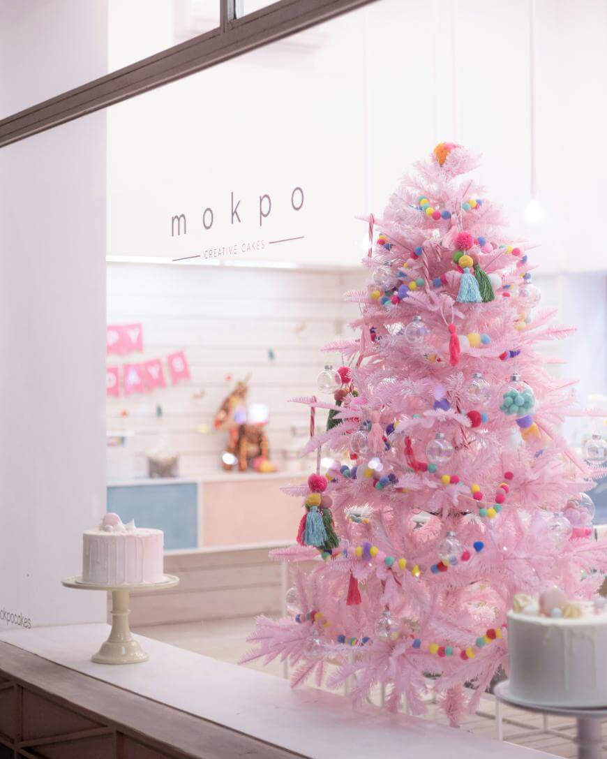 A pastel pink Christmas tree decorated with colorful pom-poms and tassels stands in a bakery window. Nearby, cakes are displayed on stands, and the shop logo “mokpo creative cakes” is visible on the glass.