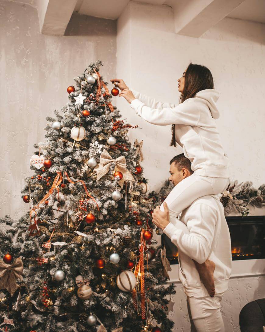 A woman in a white hoodie is sitting on a mans shoulders, reaching up to hang an ornament on a decorated Christmas tree with red and gold ornaments and ribbons. Both appear happy and festive.
