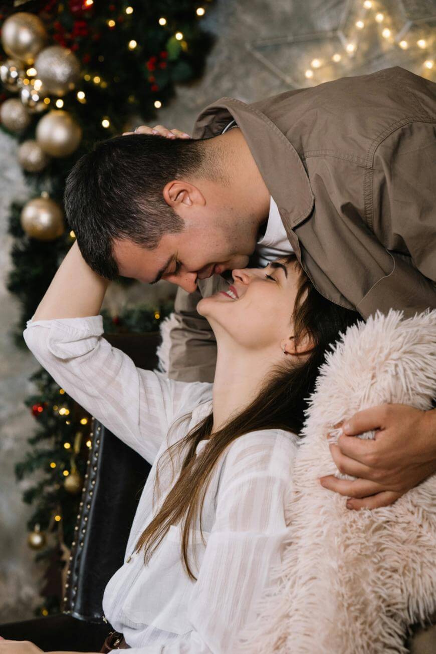 A couple shares a loving moment indoors by a decorated Christmas tree. The man leans down, smiling, while the woman reclines in a chair, smiling up at him, her arm resting on a fluffy pillow.