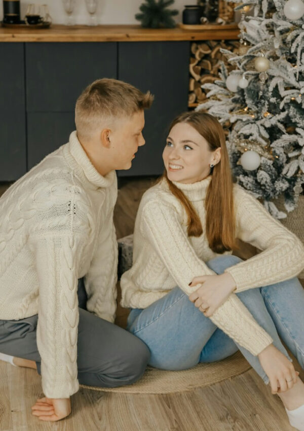 A young couple in matching cream sweaters and jeans sit on the floor by a decorated Christmas tree, smiling at each other in a cozy, festive room with wood accents.