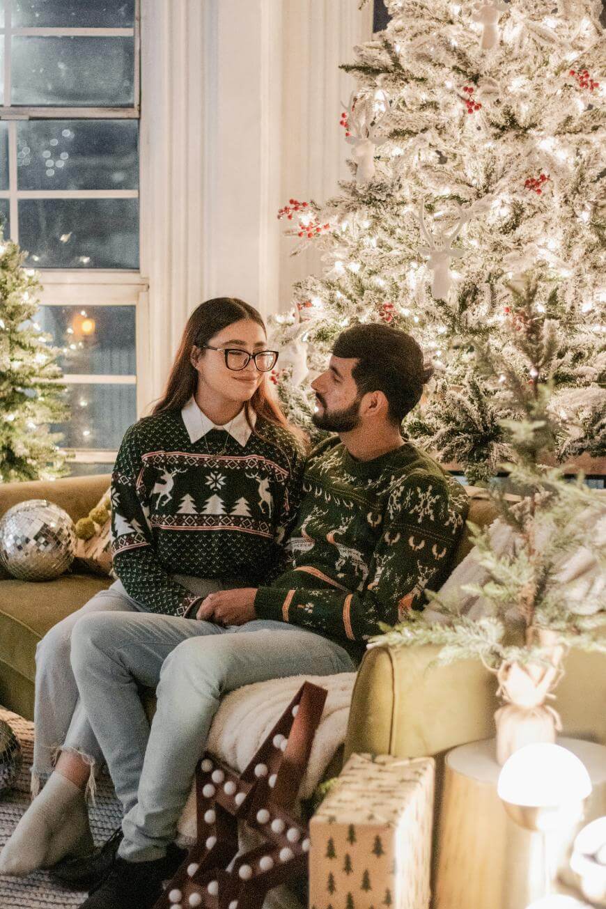 A couple wearing festive sweaters sits together on a couch by a decorated Christmas tree, surrounded by wrapped gifts and holiday lights, sharing a cozy, joyful moment.