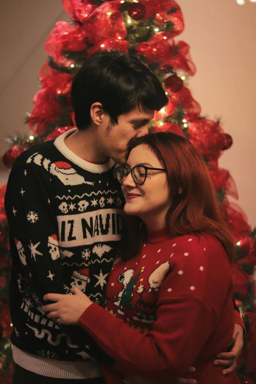 A couple in festive sweaters embraces in front of a red Christmas tree. The man kisses the woman’s forehead, and both look content and cozy, celebrating the holiday season together.
