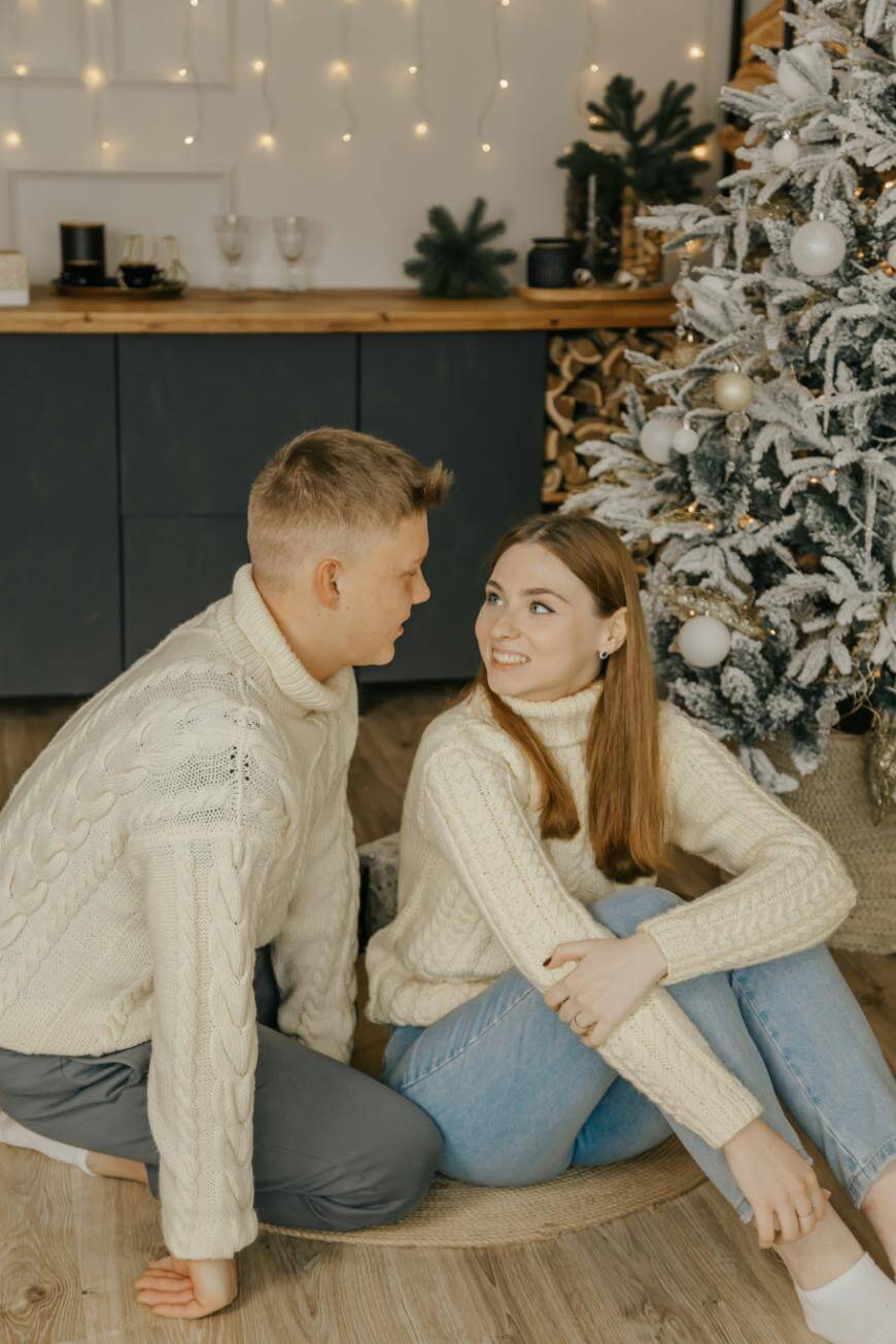 A couple in cozy white sweaters sits by a decorated Christmas tree indoors. The woman is sitting on the floor smiling at the man, who is kneeling beside her. The room is warmly lit with string lights and festive decor.