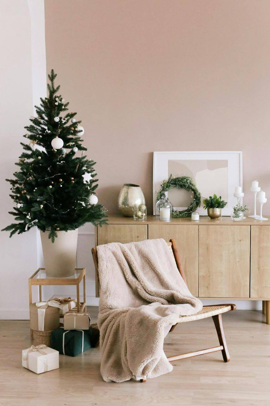 A cozy living room with a decorated Christmas tree, wrapped gifts, a wooden chair draped with a beige blanket, and a sideboard displaying candles, wreaths, and festive decor.