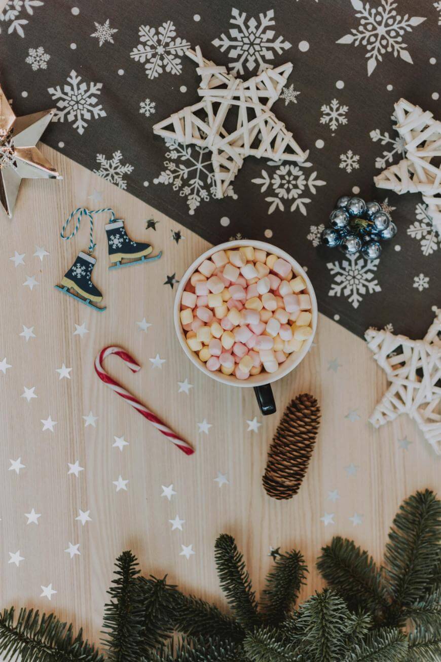 A mug of hot chocolate topped with mini marshmallows sits on a wooden table decorated with snowflake and star ornaments, a candy cane, pine cone, fir branches, and festive decorations.