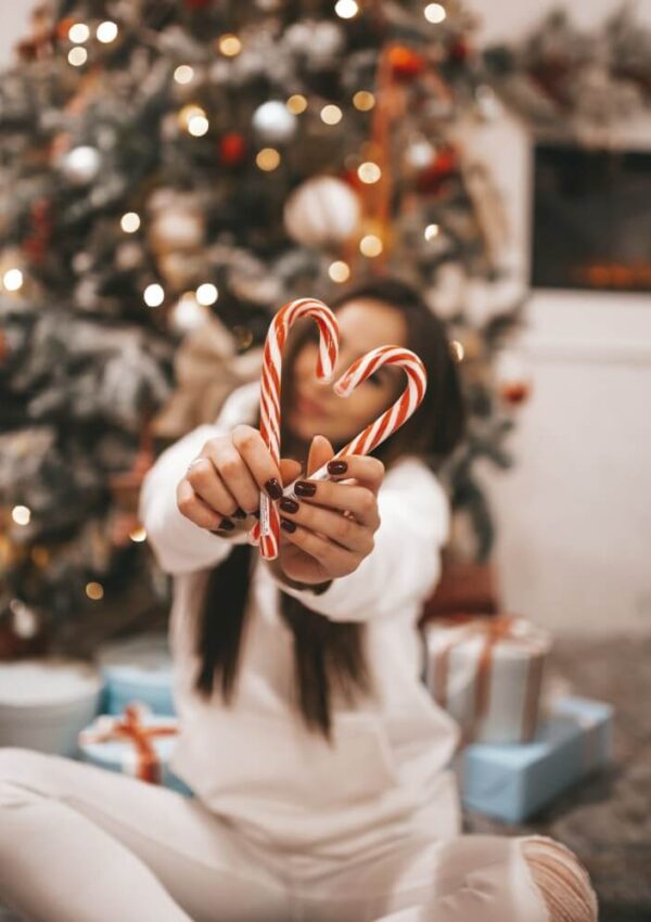 A person in white sits in front of a decorated Christmas tree, holding two candy canes shaped like a heart. Wrapped presents are on the floor, and warm lights decorate the tree in the background.